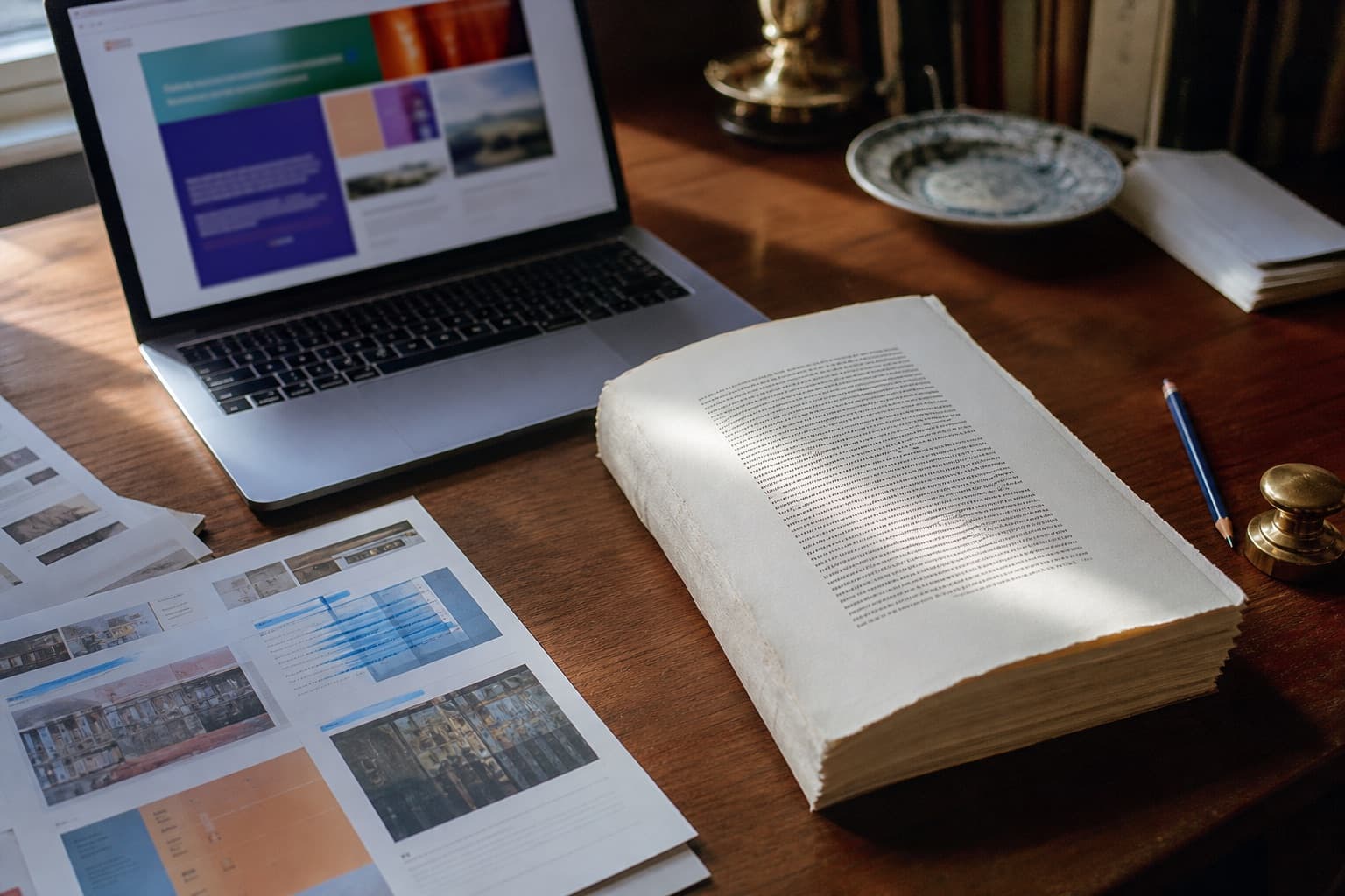Overhead of a research desk: a laptop showing a website beside an unbound cream manuscript, blue pencil, and printed pages.